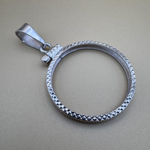 Puede incluir: Un colgante de moneda de plata con un patr&oacute;n texturizado en forma de diamante alrededor del borde. El colgante tiene una abertura con bisagras para sujetar una moneda.
