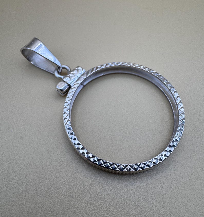 Puede incluir: Un colgante de moneda de plata con un patr&oacute;n texturizado en forma de diamante alrededor del borde. El colgante tiene una abertura con bisagras para sujetar una moneda.