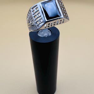 May include: A silver-toned ring with a large, rectangular black gemstone. The ring has a detailed, openwork design on the sides. The ring is displayed on a black cylindrical stand against a neutral background.