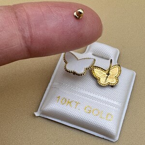 Puede incluir: Un par de pendientes de bot&oacute;n con forma de mariposa, incrustaciones blancas y bordes festoneados dorados, presentados en una tarjeta blanca con el texto 10KT. GOLD. Un pendiente muestra el frente y el otro la parte trasera con el poste.