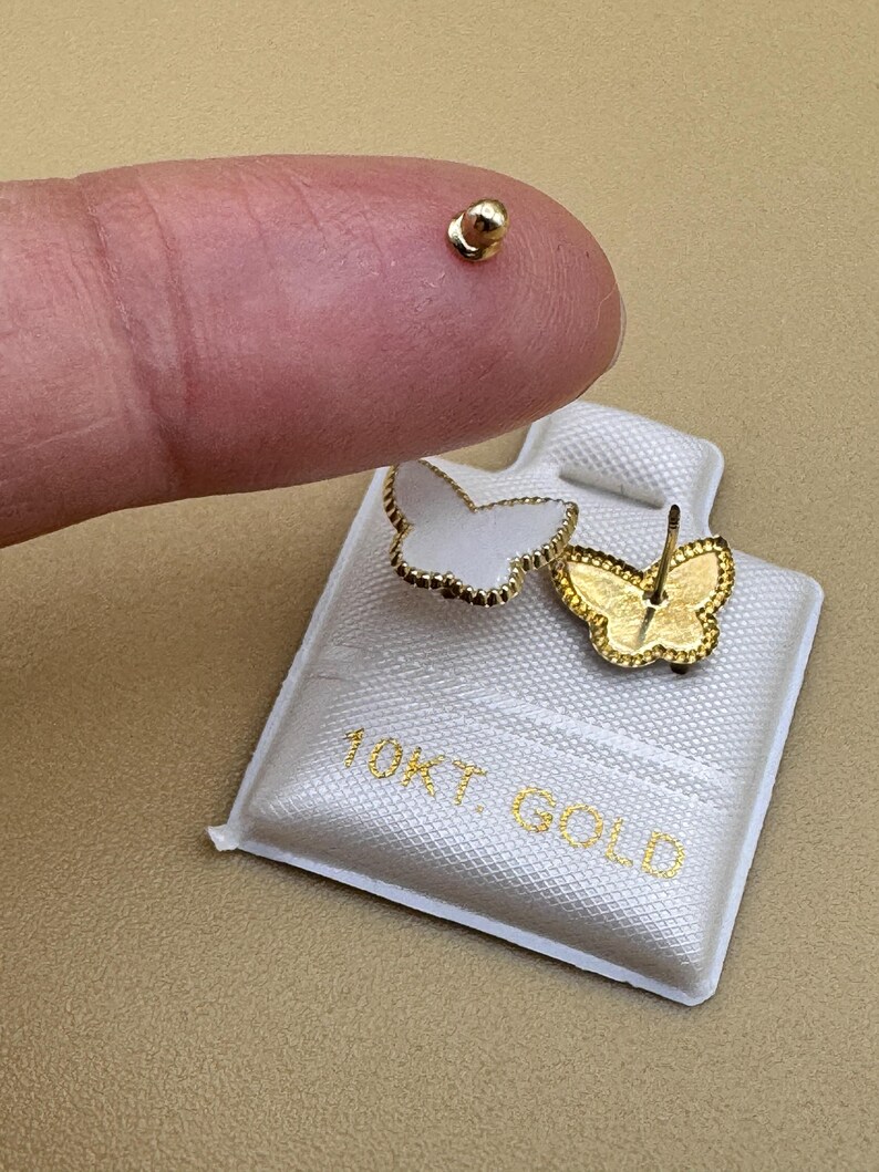 Puede incluir: Un par de pendientes de bot&oacute;n con forma de mariposa, incrustaciones blancas y bordes festoneados dorados, presentados en una tarjeta blanca con el texto 10KT. GOLD. Un pendiente muestra el frente y el otro la parte trasera con el poste.