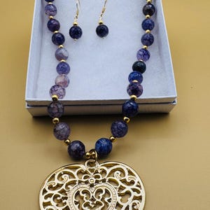 May include: A necklace and earring set featuring a beaded necklace with purple and blue beads, gold accents, and a large gold heart pendant. The earrings have matching blue beads. The jewelry is displayed in a white box.