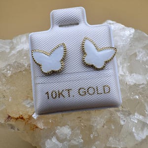 Puede incluir: Un par de pendientes de bot&oacute;n con forma de mariposa, con centros de esmalte blanco y bordes festoneados en tono dorado. Los pendientes est&aacute;n montados en una tarjeta blanca con el texto 10KT GOLD impreso en letras doradas en el frente.