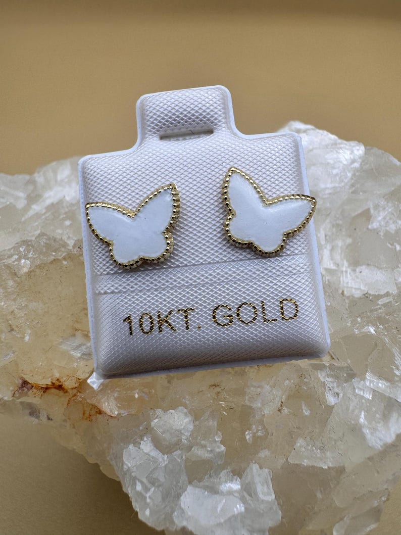 Puede incluir: Un par de pendientes de bot&oacute;n con forma de mariposa, con centros de esmalte blanco y bordes festoneados en tono dorado. Los pendientes est&aacute;n montados en una tarjeta blanca con el texto 10KT GOLD impreso en letras doradas en el frente.