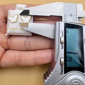 Puede incluir: Un par de pendientes en forma de mariposa blanca con bordes dorados sobre una tarjeta blanca con la etiqueta 10KT GOLD. Un calibrador digital mide el ancho de un pendiente en 7,39 mm. Los pendientes tienen un interior blanco y borde dorado.
