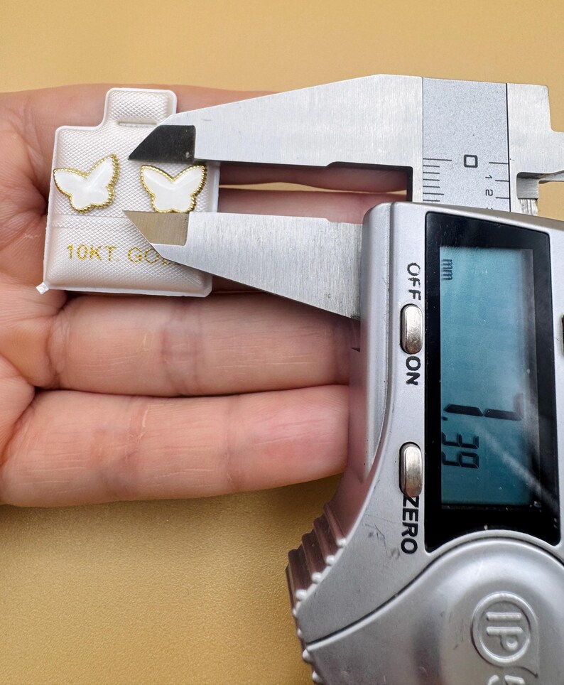 Puede incluir: Un par de pendientes en forma de mariposa blanca con bordes dorados sobre una tarjeta blanca con la etiqueta 10KT GOLD. Un calibrador digital mide el ancho de un pendiente en 7,39 mm. Los pendientes tienen un interior blanco y borde dorado.