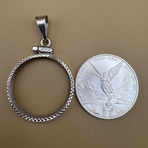 Puede incluir: Pendiente de plata con un marco circular y un cierre con bisagra. El pendiente est&aacute; dise&ntilde;ado para contener una moneda de plata con la imagen del &Aacute;ngel de la Independencia, el texto "PLATA PURA" y "2015 LEY .999".