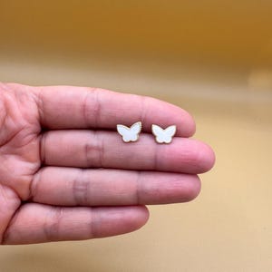 Puede incluir: Un par de pendientes de bot&oacute;n con forma de mariposa. Cada pendiente presenta un centro de esmalte blanco rodeado por un borde fino de metal dorado texturizado. El dise&ntilde;o de mariposa es sim&eacute;trico con detalles delicados en las alas y un acabado liso.