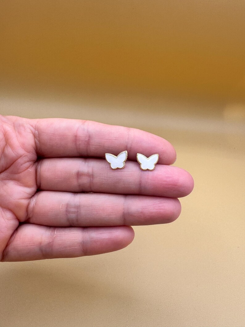 Puede incluir: Un par de pendientes de bot&oacute;n con forma de mariposa. Cada pendiente presenta un centro de esmalte blanco rodeado por un borde fino de metal dorado texturizado. El dise&ntilde;o de mariposa es sim&eacute;trico con detalles delicados en las alas y un acabado liso.
