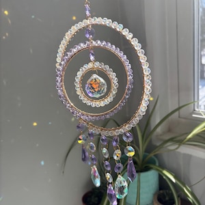 May include: A decorative suncatcher with a gold-colored metal frame and clear and purple crystal beads. The suncatcher has a spiral design with dangling crystal pendants. The crystals reflect light, creating a rainbow effect.