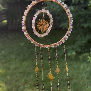 May include: A decorative hanging ornament featuring a large gold-toned wire hoop wrapped in light purple and clear crystal chips. A smaller inner hoop contains a gold sun face charm. Five gold chains with dark purple beads and sun charms hang below.