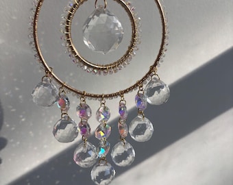 May include: A decorative suncatcher with a gold-colored metal frame. The suncatcher features two concentric circles adorned with clear crystals and hanging crystal pendants. The crystals reflect light, creating a rainbow effect.