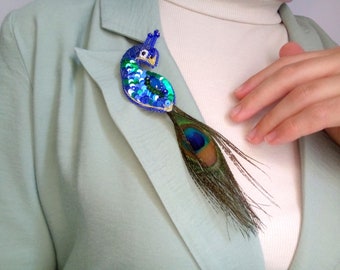 May include: A peacock brooch featuring a body crafted from blue, green, and turquoise sequins and beads, with a long, natural iridescent peacock feather tail extending downwards. The bird design includes a small beak and eye detail.