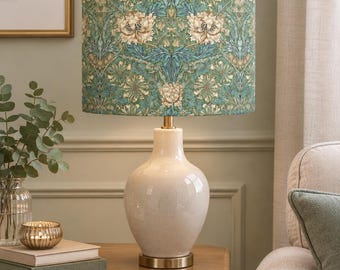 May include: A table lamp featuring a cylindrical drum shade with a teal and cream floral botanical pattern. The base is a smooth, glossy, off-white ceramic vessel with a rounded body, supported by a circular metallic gold-toned metal stand.