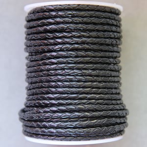 Puede incluir: Un carrete de cordón de cuero trenzado negro. El cordón está enrollado apretado y tiene un acabado liso y pulido.