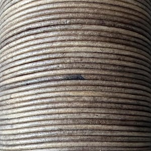 May include: Close-up of a spool of light brown leather cord. The cord is tightly wound, creating a cylindrical shape. The leather has a slightly weathered appearance, with variations in colour and texture.