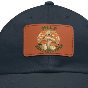 May include: Navy blue baseball cap with a rectangular patch featuring a mushroom graphic and the text "MILF MAN I LOVE FROGS". The patch is brown with orange, green, and white details. The cap is made of a soft, comfortable material.
