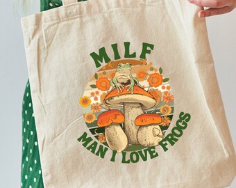 Puede incluir: Una bolsa de lona blanca con un gráfico verde, naranja y marrón de una rana sentada en un hongo. El texto "MILF MAN I LOVE FROGS" está impreso en la bolsa.