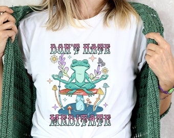 Op de afbeelding: Wit T-shirt met de tekst "DON'T HATE MEDITATE" in een retro lettertype. De afbeelding toont een mediterende kikker op een paddenstoel, omringd door bloemen en paddenstoelen. Het shirt wordt gecombineerd met een spijkerbroek en een groen vest.