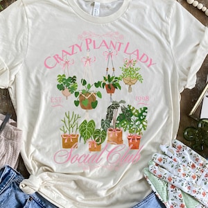 Może przedstawiać: Biały T-shirt z różowym nadrukiem, na którym widnieje napis "Crazy Plant Lady Est. 1998 Social Club". Nadruk przedstawia kolekcję roślin doniczkowych z różowymi kokardkami.