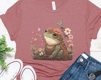 Pode incluir: Camiseta rosa empoeirado com uma ilustração caprichosa de um sapo adornado com uma coroa e capa floral, rodeado por flores e borboletas. A camiseta é combinada com tênis brancos e shorts jeans.