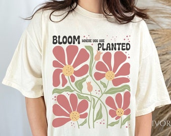 Puede incluir: Una camiseta de color crema con un diseño floral y el texto "Bloom where you are planted".
