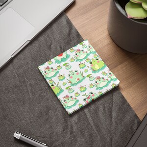 Può includere: Un blocco note bianco con un motivo ripetuto di rane verdi che indossano corone e siedono su ninfea. Le rane sono circondate da foglie verdi e fiori rosa.