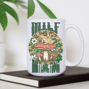 Puede incluir: Taza de cerámica blanca con una rana verde sentada en un hongo rojo y blanco. La taza tiene el texto "M.I.L.F. More Than I Love Frogs" en letras verdes y marrones.