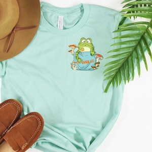 May include: A light blue t-shirt with a green frog sitting in a pocket with mushrooms and the text "Over at" printed on the pocket.