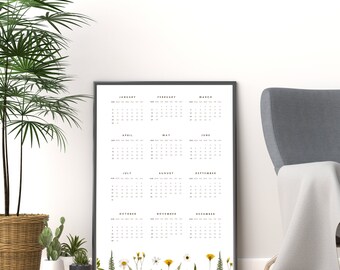 May include: A black framed calendar with a white background and black text. The calendar shows the months of the year, January through December. The calendar is hanging on a white wall with a gray armchair and a potted plant in the foreground.