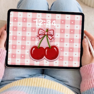Peut inclure: Une tablette affiche un motif vichy rose avec des cœurs blancs et un motif de cerises. L'heure 12h34 est visible à l'écran. La tablette est tenue par une personne portant un pull aux couleurs pastel.