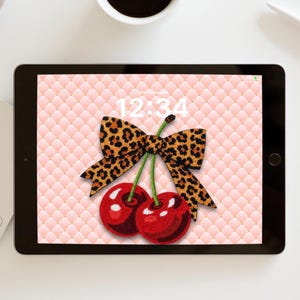Puede incluir: Una tableta digital muestra un fondo de pantalla con un lazo con estampado de leopardo y dos cerezas rojas. El fondo es un patrón acolchado rosa, y la hora 12:34 es visible. La tableta está sobre una superficie blanca.