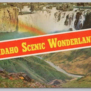 May include: A vintage postcard featuring a landscape photograph of a waterfall with a rainbow, overlaid by a diagonal red banner with the yellow text IDAHO SCENIC WONDERLAND and white pine tree silhouettes on the right and left sides.