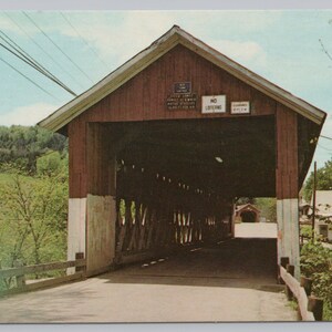 May include: A red wooden covered bridge with a gabled roof and white lower sections. The entrance features signs reading NO LOITERING and CLEARANCE 10 FT 6 IN. The interior shows a wooden truss structure, and a smaller bridge is visible in the distance.