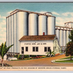 May include: Vintage postcard featuring a historic white building with the text "STARTED HERE JAN 1ST 1895", set against a backdrop of large grain storage silos. The text at the bottom reads "THE 'BIRTHPLACE' OF POST PRODUCTS IN THE SHADOW OF MODERN GRAIN STORAGE TANKS".