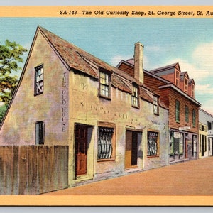 May include: Vintage postcard featuring "The Old Curiosity Shop" on St. George Street in St. Augustine, Florida. The image shows a two-story building with a weathered facade, a red-tiled roof, and the text "THE OLD HOUSE" on the side.