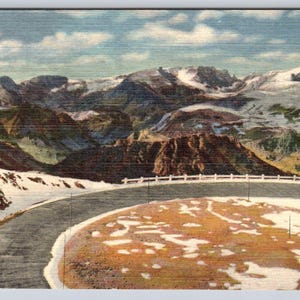 May include: Vintage postcard featuring a scenic mountain landscape with snow-capped peaks under a cloudy sky. A winding road with a white fence curves around a snow-covered area in the foreground. The image evokes a sense of travel and nature.