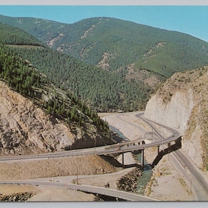 May include: A high-angle view of a multi-level highway system winding through a mountain pass. The scene features steep, rocky cliffs covered in green pine trees, a narrow river flowing beneath a concrete bridge, and a clear blue sky above.