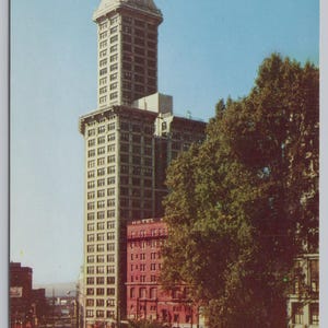 May include: Vintage postcard featuring the Smith Tower in Seattle. The tall building has a distinctive pyramid-shaped top and is surrounded by other buildings and trees. The sky is a clear blue.