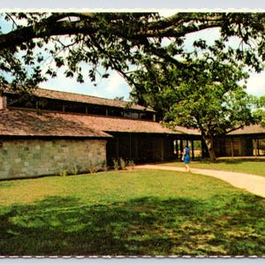 May include: A stone building with a brown roof and a walkway leading to the front entrance. The building is surrounded by green grass and trees.