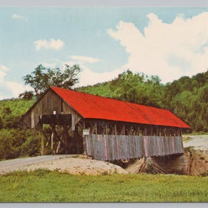May include: A vintage postcard featuring a wooden covered bridge with a bright red metal roof. The structure is supported by timber trusses and sits over a small creek, surrounded by a lush green hillside under a blue sky with white clouds.