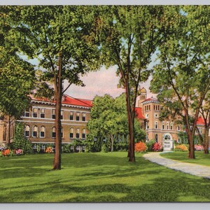 May include: Vintage postcard depicting a large brick building with a red roof, surrounded by lush green trees and a well-manicured lawn. A winding path leads to the building's entrance, with colorful flowers adding a touch of vibrancy to the scene.