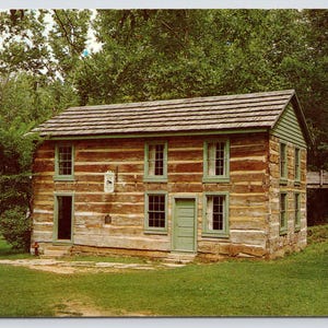 May include: A two-story log cabin with horizontal brown timber walls, a shingled roof, and light green window frames and doors. The structure features multiple rectangular windows, a small sign on the front wall, and a wooden walkway extending from the side.