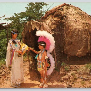 May include: Vintage postcard depicting two people in Native American attire. One person holds a ceremonial mask, while the other wears a feathered headdress and holds a bow. A traditional dwelling and wooden structure are in the background.