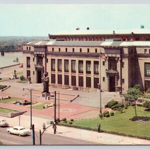 May include: A vintage postcard view of a large, multi-story stone building with a flat roof, featuring a central plaza with a statue, a fountain, and green lawns. A bridge spans the river in the background, while vintage cars drive on the street.