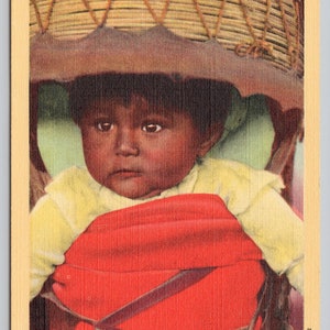 May include: Vintage postcard featuring an Apache Indian papoose. The image shows a young child in a red carrier with a woven basket overhead. The child is wearing a yellow shirt. The postcard has the text "An Apache Indian Papoose" at the top.