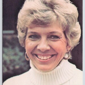 May include: A woman with short, blonde hair smiles while wearing a white turtleneck sweater.