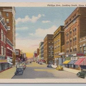 May include: Vintage postcard depicting Phillips Ave. in Sioux Falls, S.D. The image shows a street lined with brick buildings, shops, and vintage cars. The sky is a mix of blue and orange, suggesting either sunrise or sunset.