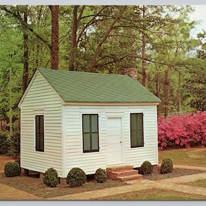 May include: A vintage postcard featuring a small, white, wooden building with a green roof and dark green shutters. The building has a white door and is surrounded by greenery, including trees and bushes. A brick pathway leads to the building.