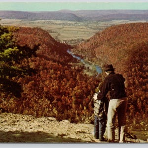 May include: A vintage postcard showing a scenic overlook of a valley with dense autumn foliage in shades of orange and brown. A winding river flows through the center, while two figures stand on a rocky cliff edge in the foreground looking out.